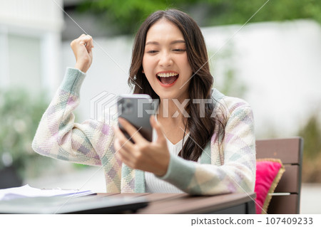 Millennial asian young woman looking mobile phone laughing with good news or discount voucher for shopping online at home.Happy and cheerful woman looking on cellphone app read message feel excited 107409233