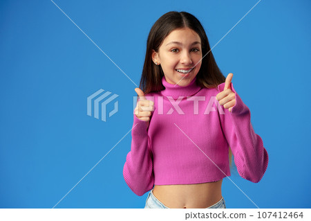 Portrait of joyful teenage girl showing thumbs up on blue background 107412464