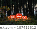 a group of carved pumpkins perched on top of a mound at the edge of a forest. 107412612