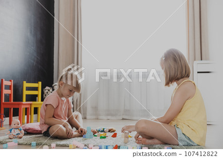 Children playing with colorful wooden toy building blocks, bricks. Kids sitting on floor at sunny white home bedroom or kindergarten, daycare playroom. Funny educational eco-friendly game for child 107412822