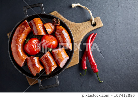 Fatty pork sausage fried with whole tomatoes in a cast iron frying pan on a wooden cutting board with cutlery on a black concrete table. Top view with copy space Fatty pork sausage fried with whole tomatoes in a cast iron frying pan on a wooden cutting board with cutlery on a black concrete table. Top view with copy space 107412886