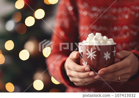 woman's hands holding a cup of hot chocolate with marshmallows with a Christmas tree in the background 107413032