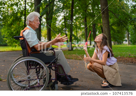 The man in a wheelchair having fun with his caregiver in the park, catching bubbles. The man in a wheelchair having fun with his caregiver in the park, catching bubbles. 107413576