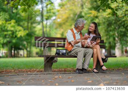 The grandfather and granddaughter are looking at photos on a tablet, sitting on a bench in a city park. 107413630