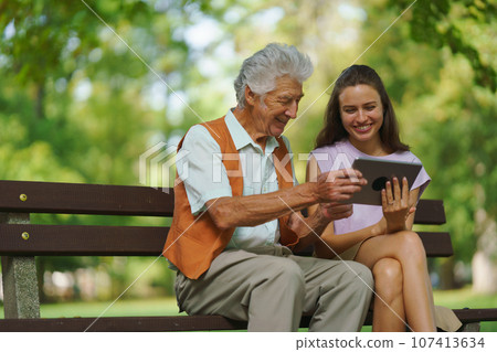 The grandfather and granddaughter are looking at photos on a tablet, sitting on a bench in a city park. 107413634