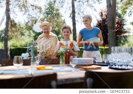 Grandmother, mother and daughter setting table for summer garden party. Grandmother, mother and daughter setting table for summer garden party. 107413731