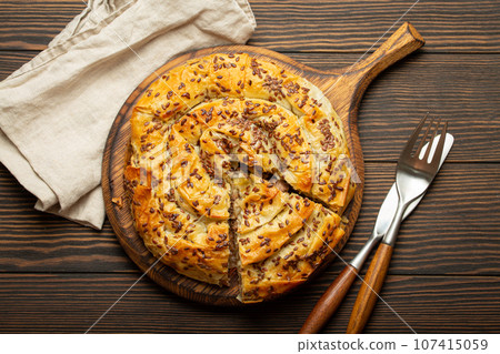 Burek with cut piece made of filo dough with filling on cutting board, dark brown wooden rustic background top view. Traditional savoury spiral pie of Balkans, Middle East and Central Asia 107415059