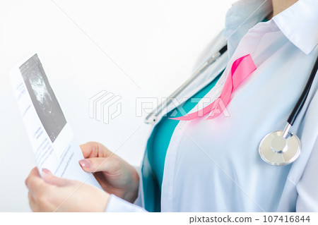 Woman doctor with pink ribbon on white medical uniform holds results of ultrasound scan in her hands. Breast cancer awareness month symbol. Woman doctor with pink ribbon on white medical uniform holds results of ultrasound scan in her hands. Breast cancer awareness month symbol. 107416844
