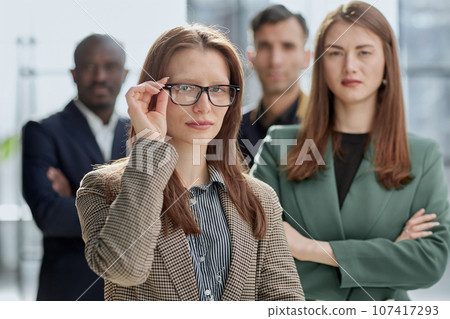 businesswoman in formal wear adjusting her glasses looking at the camera while standing in the office instead of with colleagues 107417293