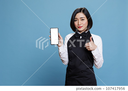 Young asian woman cafe worker presenting smartphone empty screen mockup and showing thumb up gesture. Waitress showcasing mobile phone blank touchscreen and expressing approval sign portrait Young asian woman cafe worker presenting smartphone empty screen mockup and showing thumb up gesture. Waitress showcasing mobile phone blank touchscreen and expressing approval sign portrait 107417956