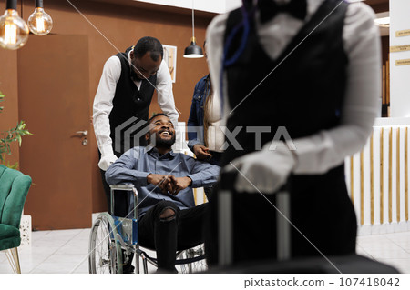 Wheelchair-friendly travel. Helpful hotel staff helping happy young male disabled guest at reception area, bellman pushing assisting man on wheelchair. Tourism and people with disabilities 107418042