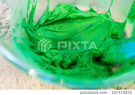 In a well-lit kitchen, an assortment of buttercream frosting in various shades of green and other colors is being meticulously mixed and prepared for the artistic decoration of cupcakes in charming In a well-lit kitchen, an assortment of buttercream frosting in various shades of green and other colors is being meticulously mixed and prepared for the artistic decoration of cupcakes in charming 107419858