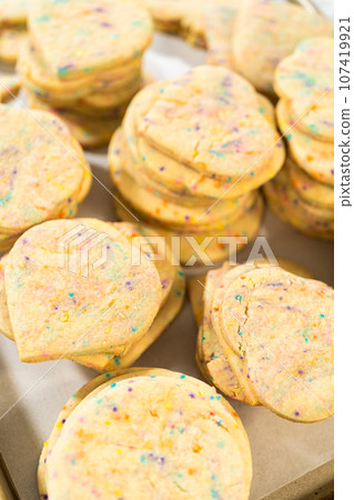 Large quantity of sprinkle-filled sugar cookies are neatly stacked on a baking tray, awaiting their packaging into the rustic brown paper boxes. 107419921