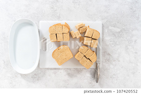 Flat lay. Cutting stale bread with a bread knife on a white cutting board to make bread pudding. 107420552