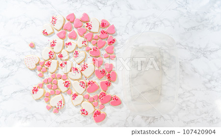 Flat lay. Storing heart-shaped sugar cookies with pink and white royal icing in a large plastic container. 107420904