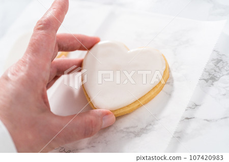 Decorating heart-shaped sugar cookies with pink and white royal icing for Valentine's Day. Decorating heart-shaped sugar cookies with pink and white royal icing for Valentine's Day. 107420983