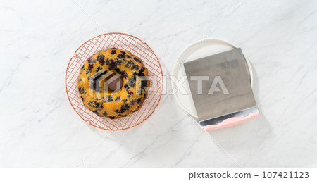 Flat lay. Transferring cooled freshly baked lemon blueberry bundt cake from the round cooling rack to the serving plate. Flat lay. Transferring cooled freshly baked lemon blueberry bundt cake from the round cooling rack to the serving plate. 107421123