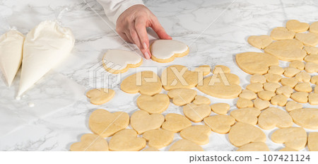 Decorating heart-shaped sugar cookies with pink and white royal icing for Valentine's Day. Decorating heart-shaped sugar cookies with pink and white royal icing for Valentine's Day. 107421124