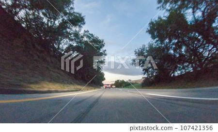 As the sun sets, driving on HWY 101 near Pismo Beach, California becomes a captivating experience with the sky painted in shades of orange and pink, casting a warm glow over the coastal landscape. 107421356