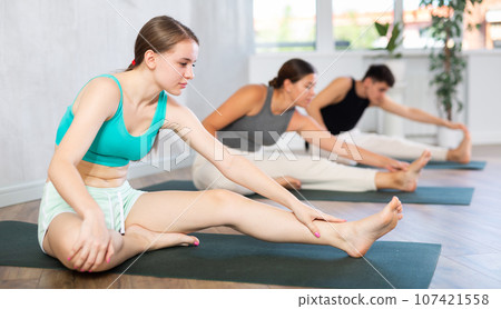 Young people doing one leg forward bend pose in class. 107421558