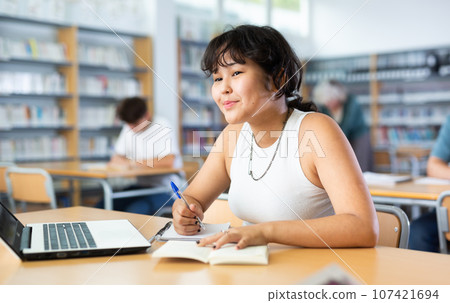 Portrait of focused teenage female pupil sitting at desk studying in library with classmates 107421694