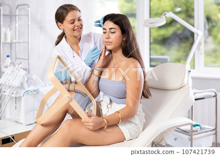 Cosmetologist explaining facial procedure to young girl looking in mirror Cosmetologist explaining facial procedure to young girl looking in mirror 107421739