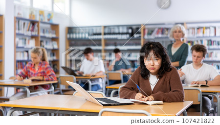 Schoolgirl studies on a laptop and writes notes in a copybook Schoolgirl studies on a laptop and writes notes in a copybook 107421822