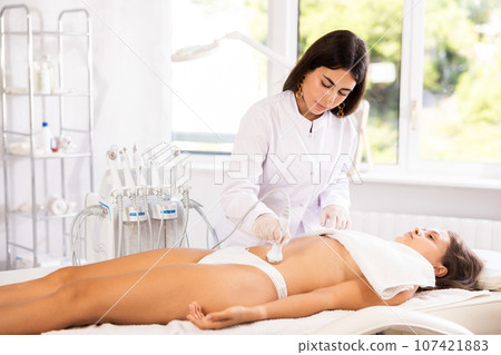 Cosmetologist performing ultrasound cavitation procedure on belly for female client Cosmetologist performing ultrasound cavitation procedure on belly for female client 107421883