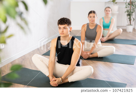In sport studio with small group of friends, young man performs butterfly pose, Baddha Konasana 107421925