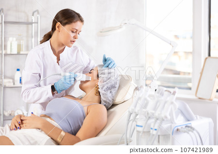 European aesthetician making facial beauty procedure with leather knife attachment to brunette girl 107421989