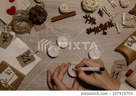 Unrecognizable young woman paint numbers on tea candles. Female making homemade advent calendar Made with your own hands step by step DIY crafts do it yourself. Preparation to christmas concept 107428703