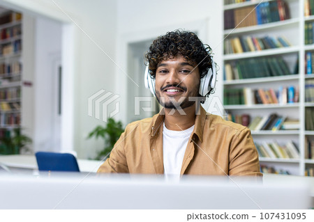 Close-up portrait of a young Indian male student in headphones studying in the library using a laptop. 107431095