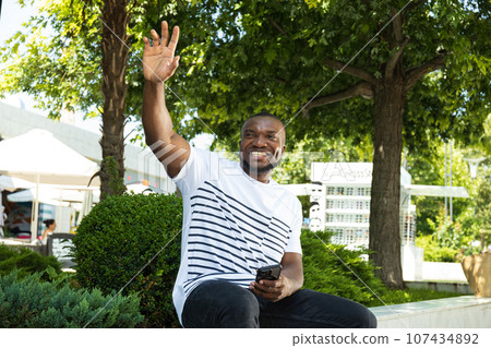 A young African-American man with a smartphone is sitting on a park bench A young African-American man with a smartphone is sitting on a park bench 107434892