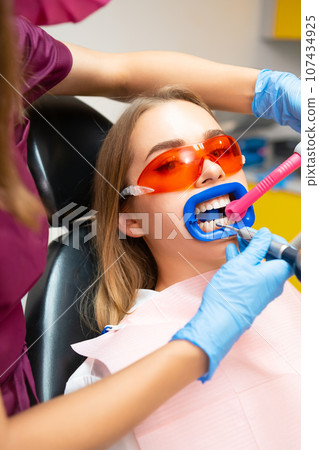 Woman wearing protective goggles from ultraviolet light sits with tube in mouth to absorb liquid. Dentist places tube in patient mouth and works on teeth Woman wearing protective goggles from ultraviolet light sits with tube in mouth to absorb liquid. Dentist places tube in patient mouth and works on teeth 107434925