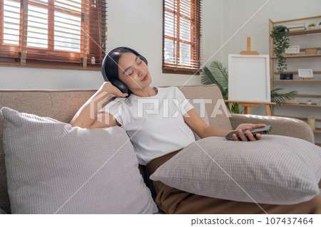 Young Asians woman listen to music on couch in living room at home. Happy Asia female using mobile smartphone, wearing headset and sitting on sofa 107437464