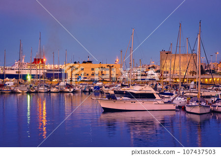 View of port area and bay in Heraklion, capital of Crete island, Greece. Ferries and cruises, fishing boats moored in harbor at sunset. Clear blue sky, deep blue waters, yachts, sailboats. Travel 107437503