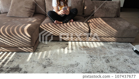 Young woman is using smartphone touching screen while robotic vacuum cleaner is cleaning carpet doing housework. Modern technology of smart cleaning. 107437677