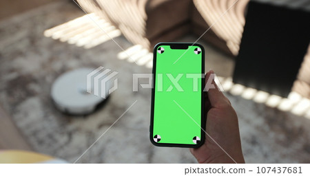 Robot vacuum cleaner performs automatic cleaning of the house at a certain time. Woman using mobile phone with green screen in living room. Green screen chroma key. Smart home concept. Robot vacuum cleaner performs automatic cleaning of the house at a certain time. Woman using mobile phone with green screen in living room. Green screen chroma key. Smart home concept. 107437681