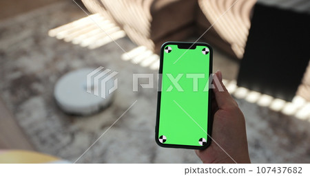 Robot vacuum cleaner performs automatic cleaning of the house at a certain time. Woman using mobile phone with green screen in living room. Green screen chroma key. Smart home concept. Robot vacuum cleaner performs automatic cleaning of the house at a certain time. Woman using mobile phone with green screen in living room. Green screen chroma key. Smart home concept. 107437682