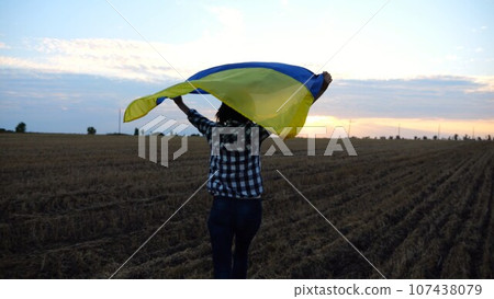 Ukrainian woman running with raised flag Ukraine above her head on wheat field at sunset. Lady jogging with national blue-yellow banner on barley meadow at sunrise. Victory against russian aggression. Ukrainian woman running with raised flag Ukraine above her head on wheat field at sunset. Lady jogging with national blue-yellow banner on barley meadow at sunrise. Victory against russian aggression. 107438079