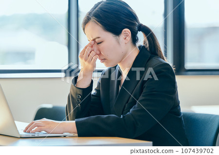 A young woman puts her hand between her eyebrows while operating a computer A young woman puts her hand between her eyebrows while operating a computer 107439702