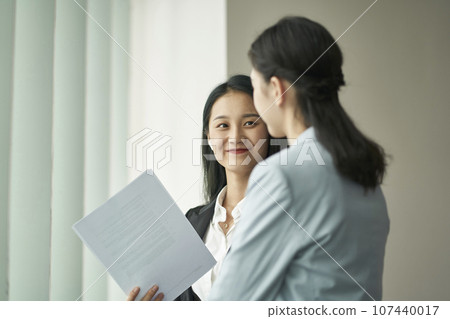 two young asian business women standing by window having a discussion in office 107440017