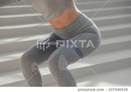 Sporty athletic woman in grey sportswear doing exercises with gymnastic elastic bands sit-ups outdoors Sporty athletic woman in grey sportswear doing exercises with gymnastic elastic bands sit-ups outdoors 107440426