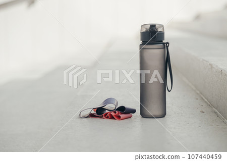 Gray sport bottle drinking water flask and gymnastic elastic bands on white stair. Hydration or thirst during fitness Gray sport bottle drinking water flask and gymnastic elastic bands on white stair. Hydration or thirst during fitness 107440459