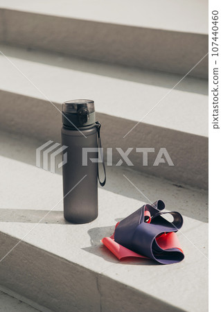 Gray sport bottle drinking water flask and gymnastic elastic bands on white stair. Hydration or thirst during fitness Gray sport bottle drinking water flask and gymnastic elastic bands on white stair. Hydration or thirst during fitness 107440460