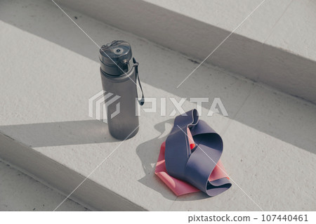 Gray sport bottle drinking water flask and gymnastic elastic bands on white stair. Hydration or thirst during fitness Gray sport bottle drinking water flask and gymnastic elastic bands on white stair. Hydration or thirst during fitness 107440461