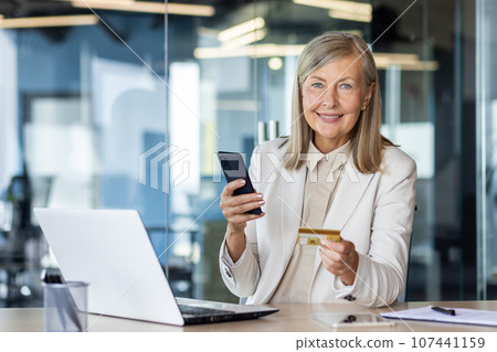 Portrait of a senior business woman working in the office using a laptop, holding a mobile phone and a credit card. Portrait of a senior business woman working in the office using a laptop, holding a mobile phone and a credit card. 107441159