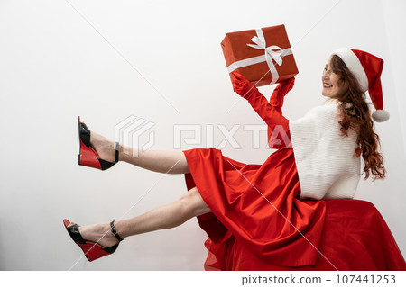 Looking enthusiastic, a woman in a Santa Claus hat and a black dress and gloves and a knitted scarf holds a special gift in her hands, preparing to open it for a winter holiday. Looking enthusiastic, a woman in a Santa Claus hat and a black dress and gloves and a knitted scarf holds a special gift in her hands, preparing to open it for a winter holiday. 107441253