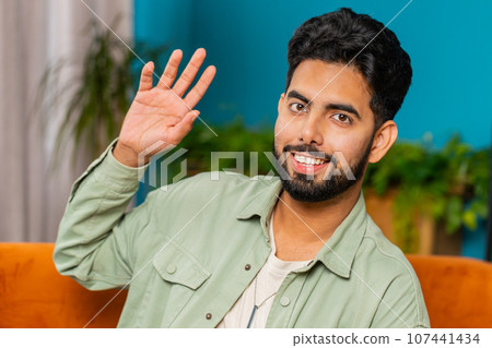 Indian man smiling friendly at camera, waving hands gesturing hello, hi, greeting at home on sofa Indian man smiling friendly at camera, waving hands gesturing hello, hi, greeting at home on sofa 107441434