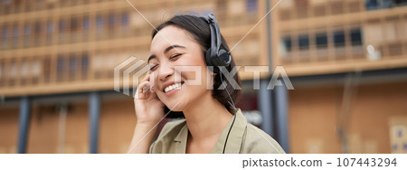 Portrait of beautiful asian woman in headphones, listening music on street of city centre, smiling happily Portrait of beautiful asian woman in headphones, listening music on street of city centre, smiling happily 107443294